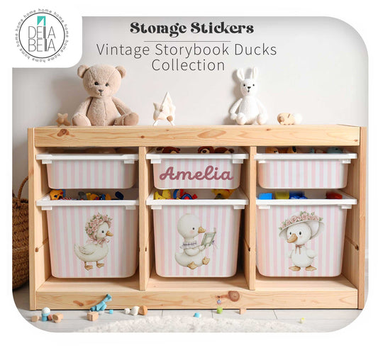 A trofast wooden storage unit with six white storage bins decorated with custom name and pink stripes and duck illustrations, topped with stuffed toys. The bins are arranged on a light wood floor in a nursery-like setting.